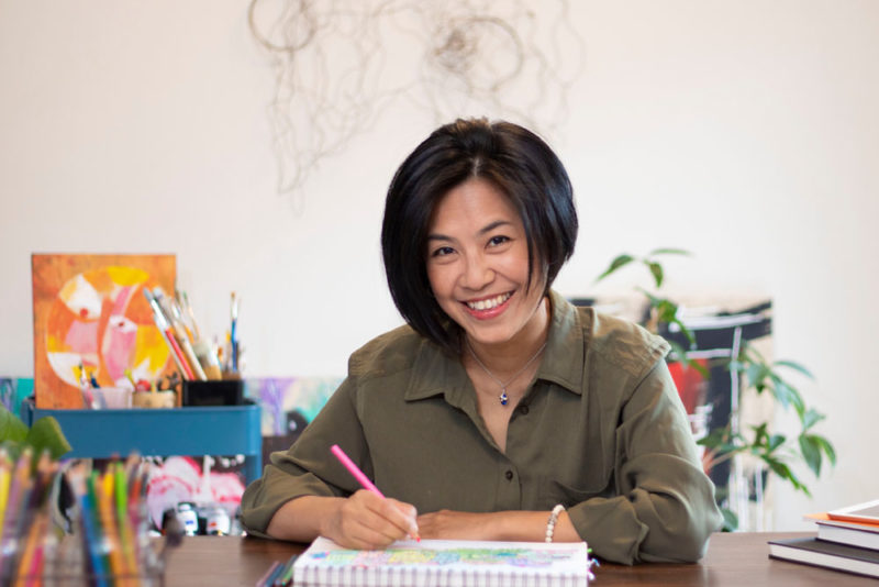 An image of illustrator, Jeni Chen, as she writes on a piece of paper with drawing supplies beside her.